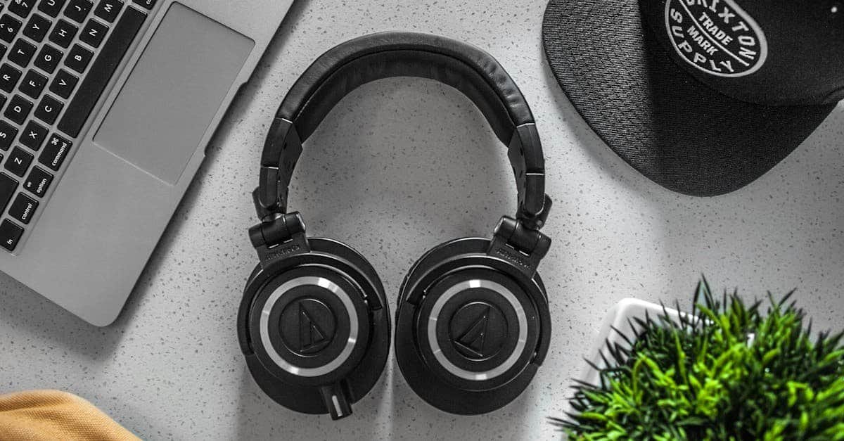 Black wireless over-ear headphones on desk with laptop and cap.