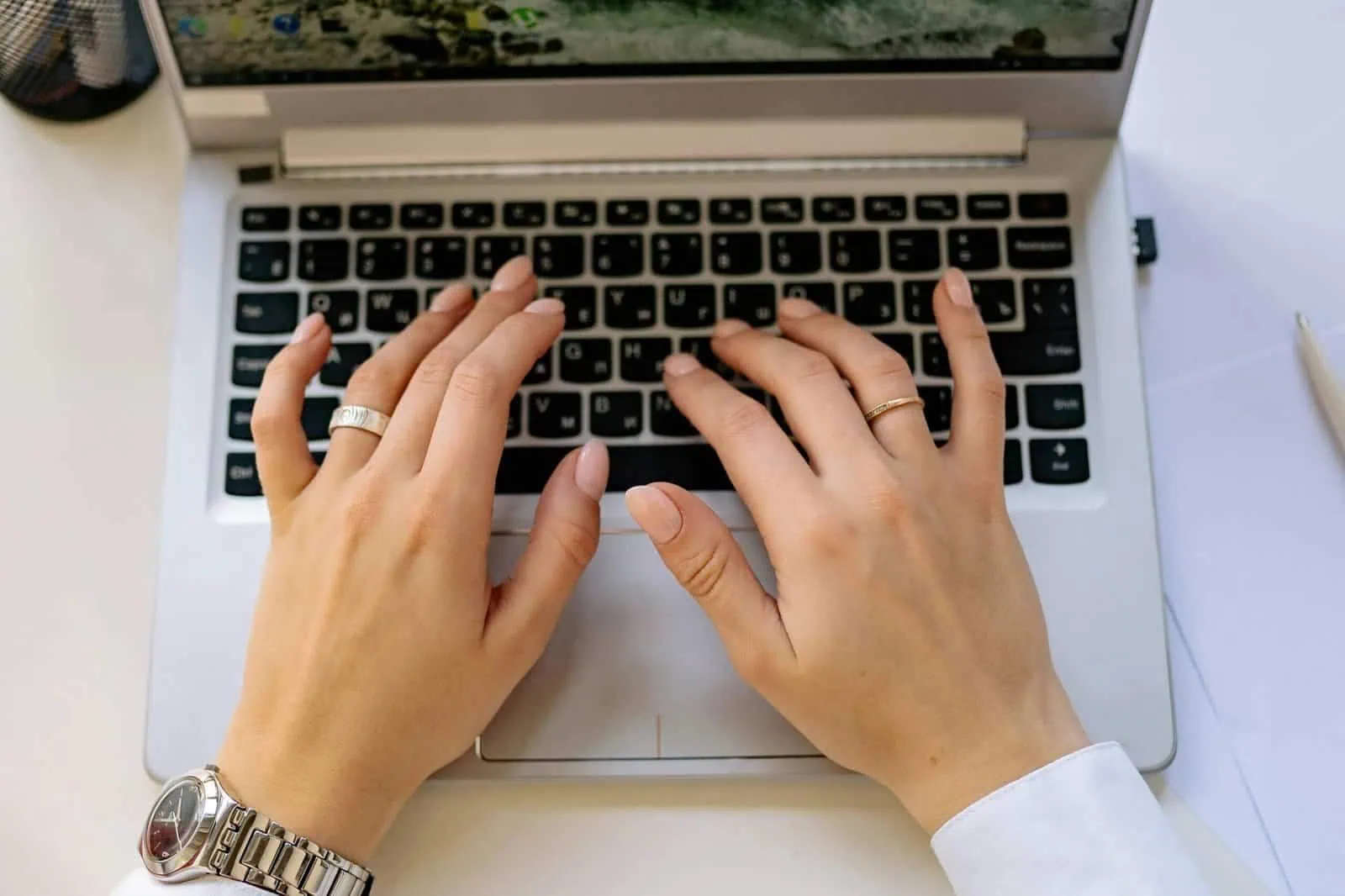 Laptop typing hands with a watch and rings on a white desk.