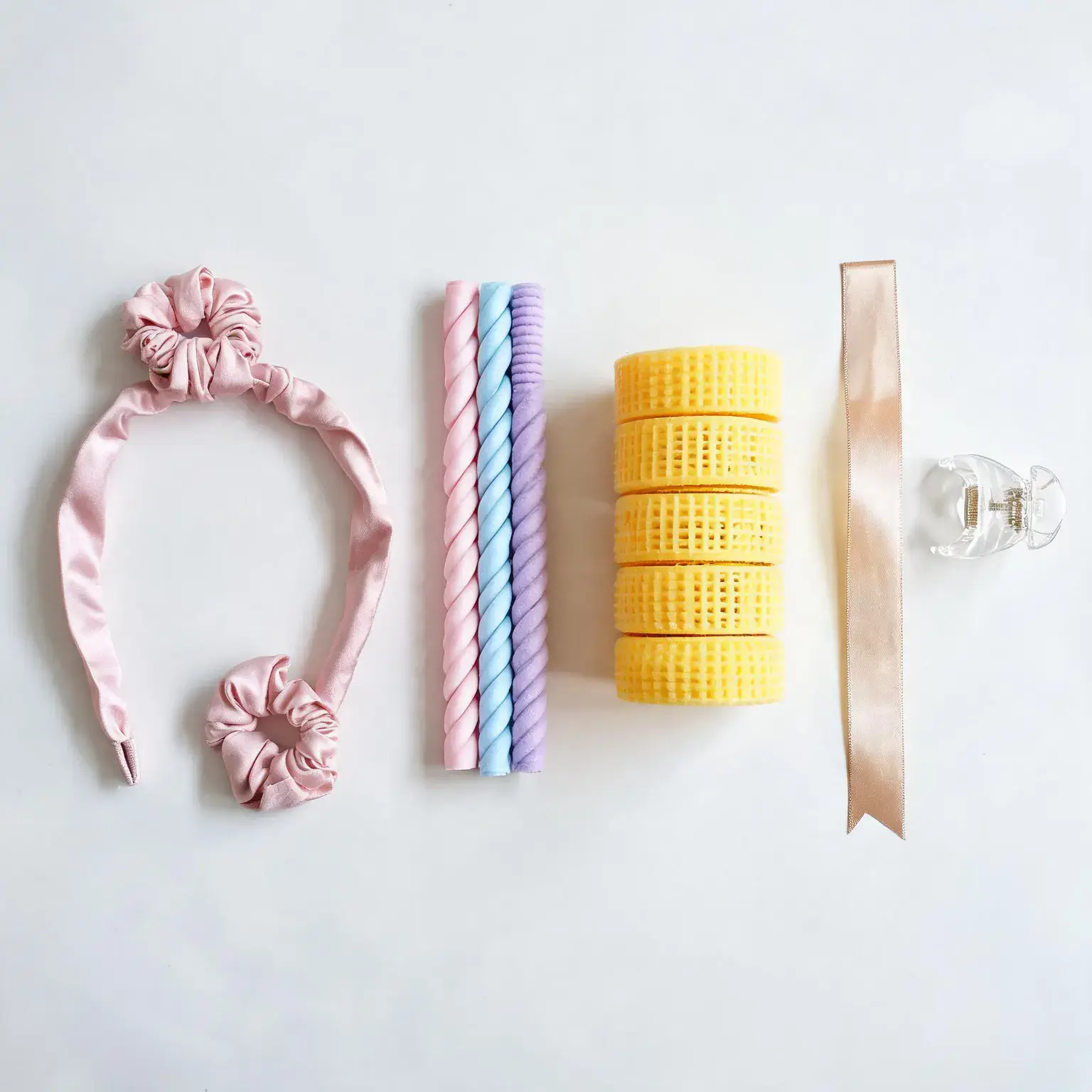 Four heatless curler formats compared side by side: a pink satin curling rod headband with two scrunchies, three pastel foam flexi rods, a stack of yellow self-grip velcro rollers, and a champagne silk ribbon wrap with a clear claw clip on a white studio background