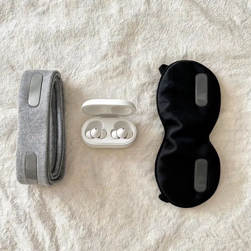 Three styles of sleep headphones compared on cream linen: a folded fabric wireless bluetooth headband, tiny silicone sleeping earbuds in an open charging case, and a black contoured sleep mask with built-in flat speakers